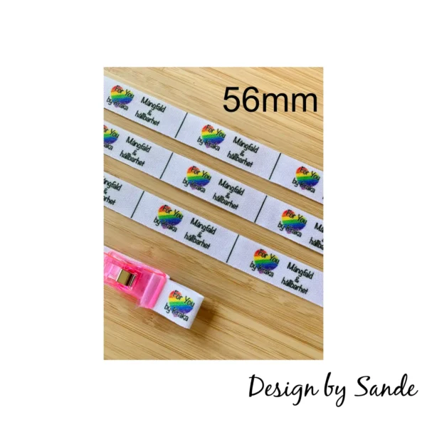 Foto av 56 mm långa etiketter på 16 mm brett vävt polyesterband med regnbågsfärgad logga och texten "Mångfald & hållbarhet". En etikett hålls uppvikt med rosa klämma.